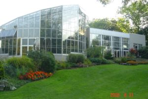 Tall rounded glass greenhouse witht eh front lined with gardens, a mix of bushes and brightly coloured flowers. 